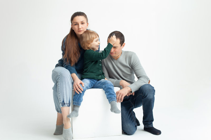A woman and man sit with a young boy on a white cube; the boy is touching the mans forehead. All three, dressed casually, have neutral expressions against a plain white background, suggesting a quiet moment possibly related to finance or divorce.