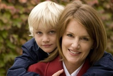 A woman smiles at the camera while a young blond boy hugs her from behind. Both are outdoors with blurred foliage in the background, portraying the warmth and trust often shared between a financial advisor and her clients.