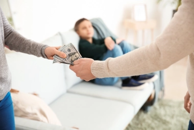 Two people exchange dollar bills—discussing child support while a child sits on a couch in the background. A divorce financial planner can help you get the financial support you deserve.