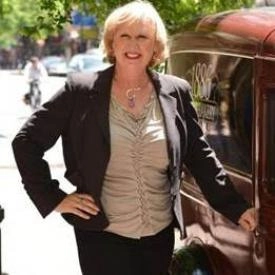 A woman with blonde hair wearing a black blazer and light-colored blouse stands outdoors, smiling and resting one hand on a maroon vehicle—embodying the confidence of a skilled divorce financial planner.