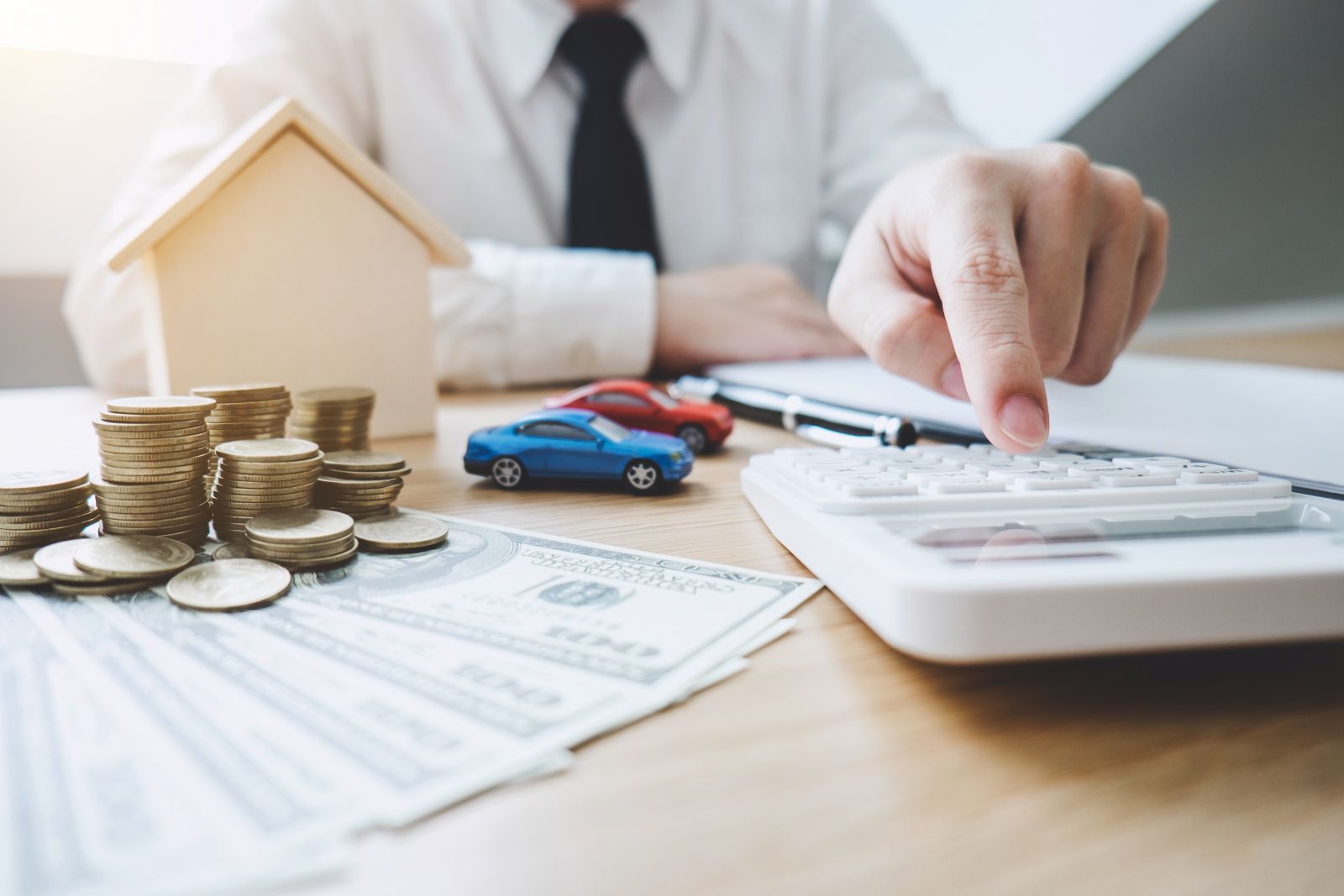 A person uses a calculator at a desk with model cars, a wooden house, coin stacks, and US dollar bills, suggesting finance planning or budgeting.