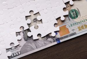 A partially completed white jigsaw puzzle covers a U.S. one hundred dollar bill on a dark wooden surface, symbolizing how a divorce financial planner can help piece together your finances during divorce.