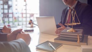Two people in suits sit across a desk from a professional with a laptop; a gavel, scales of justice, and notebooks are on the desk, suggesting a divorce or financial consultation with an advisor.