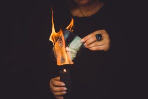 A person holding a lit candle burns a photograph, the flames consuming the photo against a dark background—symbolizing money lost or memories fading after divorce.