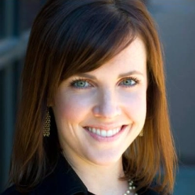 A woman with straight brown hair and blue eyes smiles at the camera, wearing a black top, pearl necklace, and gold earrings—her confident presence reflects her expertise as a financial advisor.