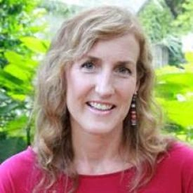 A woman with wavy, light brown hair and earrings smiles at the camera outdoors, with green foliage in the background—radiating the confidence of a financial advisor ready to help you manage your money.