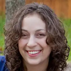 A person with curly brown hair smiling outdoors, perhaps a financial advisor enjoying a break, with a tree and wooden fence visible in the background.