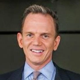 A man wearing a suit and tie smiles at the camera in front of a dark, blurred background, embodying the confidence of a financial advisor ready to help navigate life’s money matters.