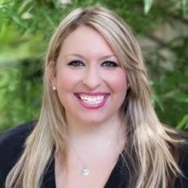 A woman with long blonde hair, a black top, and a necklace smiles at the camera, standing outdoors with green foliage behind her—a confident divorce financial planner ready to guide you through lifes transitions.
