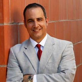 A divorce financial planner in a light gray suit and red tie stands with arms crossed, leaning against a brick wall and smiling at the camera.