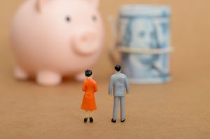 Two miniature figurines face a pink piggy bank and a rolled-up bill secured with a rubber band, symbolizing dividing Retirement accounts in a divorce