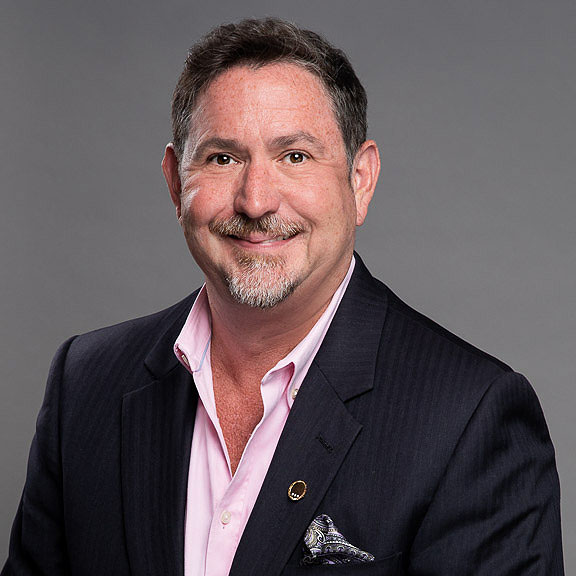 A middle-aged man with short dark hair and a goatee, wearing a dark suit jacket, light pink shirt, and pocket square, poses against a plain gray background.