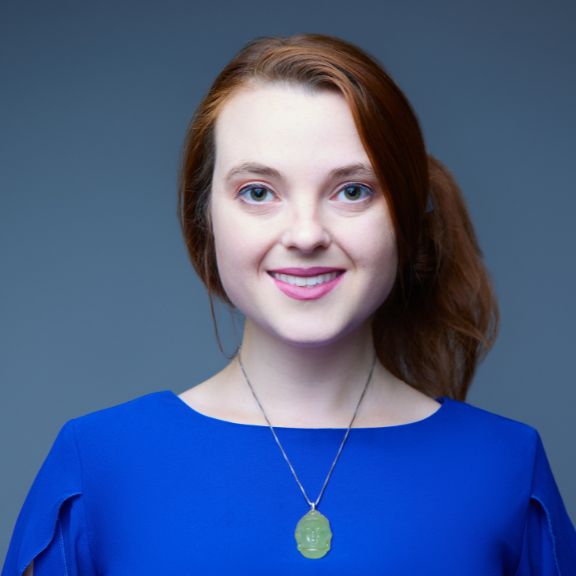 Woman with long red hair in a ponytail, wearing a bright blue top and a pendant necklace, smiling at the camera.