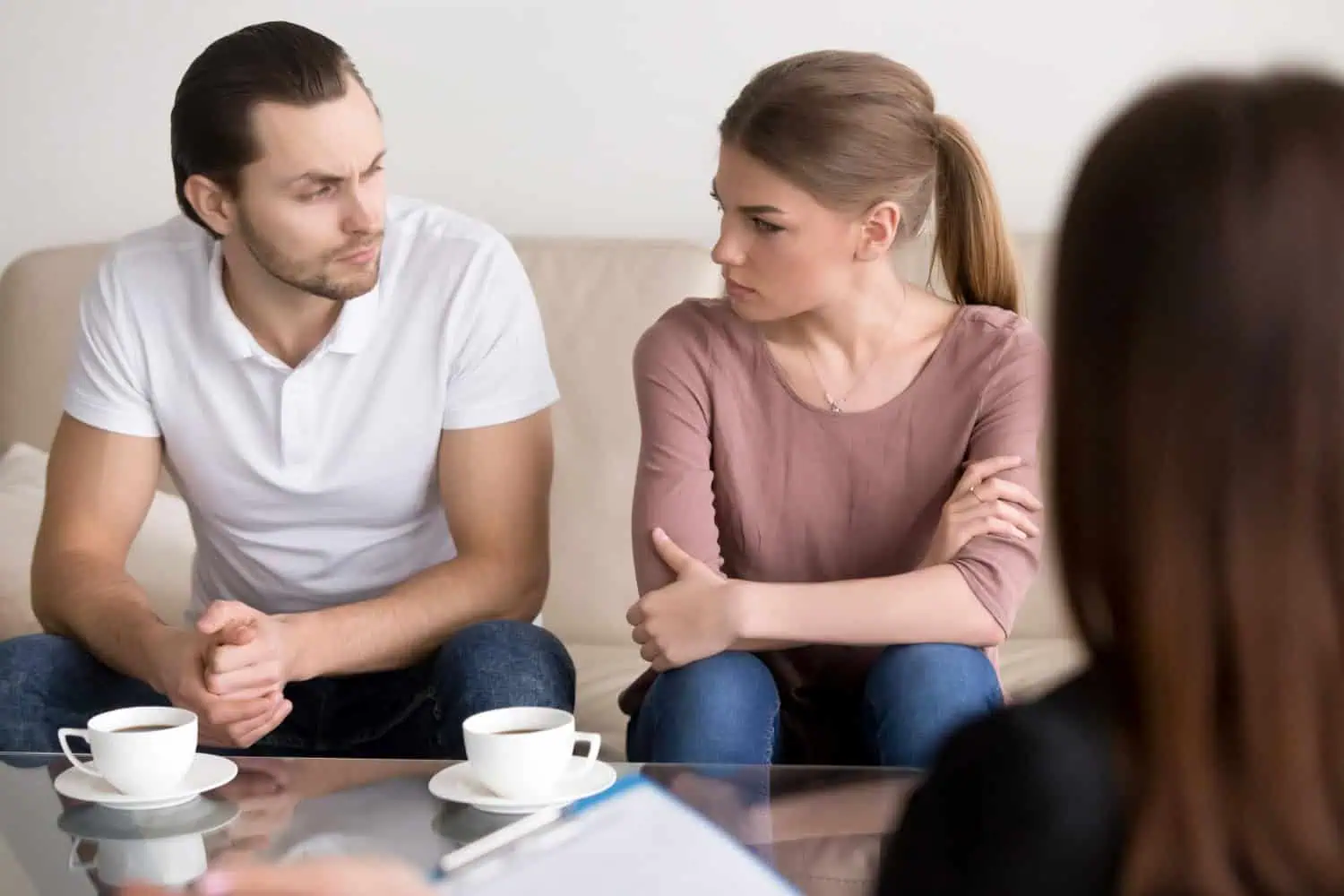 A couple sits on a sofa, looking at each other, during a therapy session with a professional.